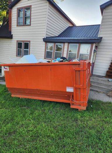 An orange dumpster on a grassy lawn next to a two-story house with siding and a dark metal roof.
