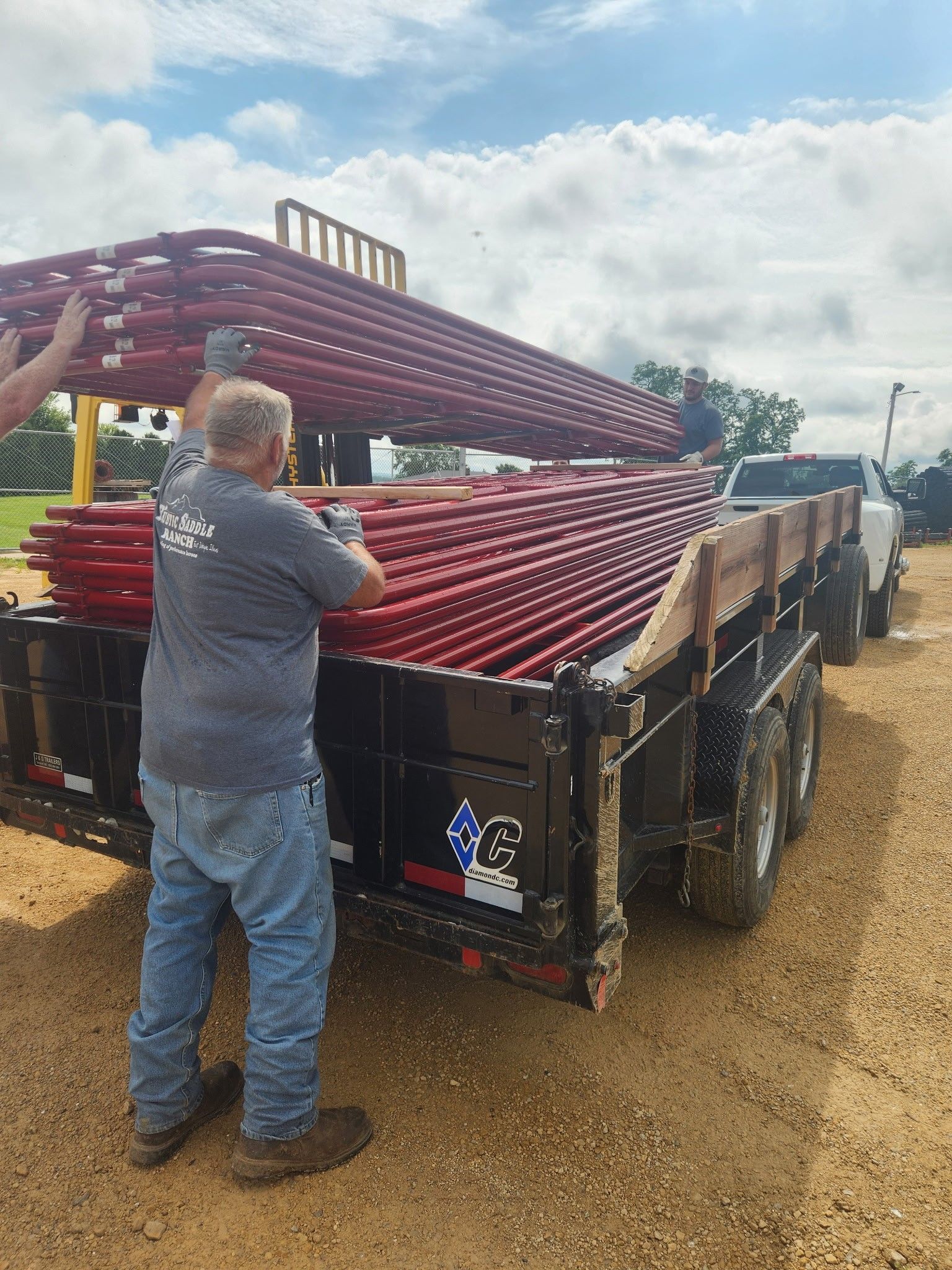 Two people loading red metal pipes onto a trailer outdoors under a cloudy sky.