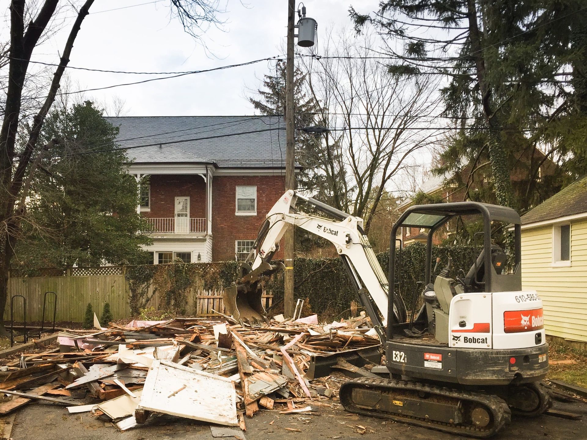 Bobcat excavator demolishing a structure, debris scattered on the ground. A brick house is in the background.