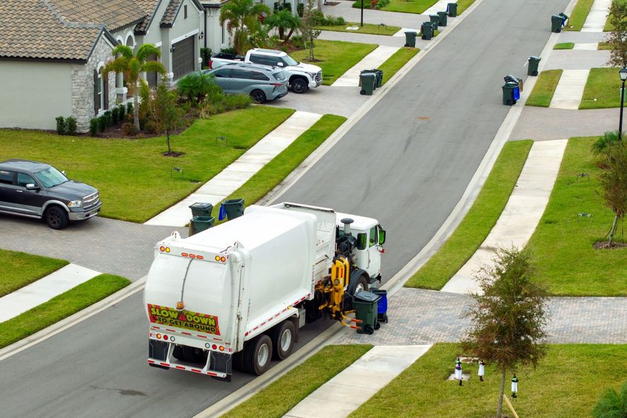 Garbage truck collecting bins on a residential street lined with houses and green lawns.