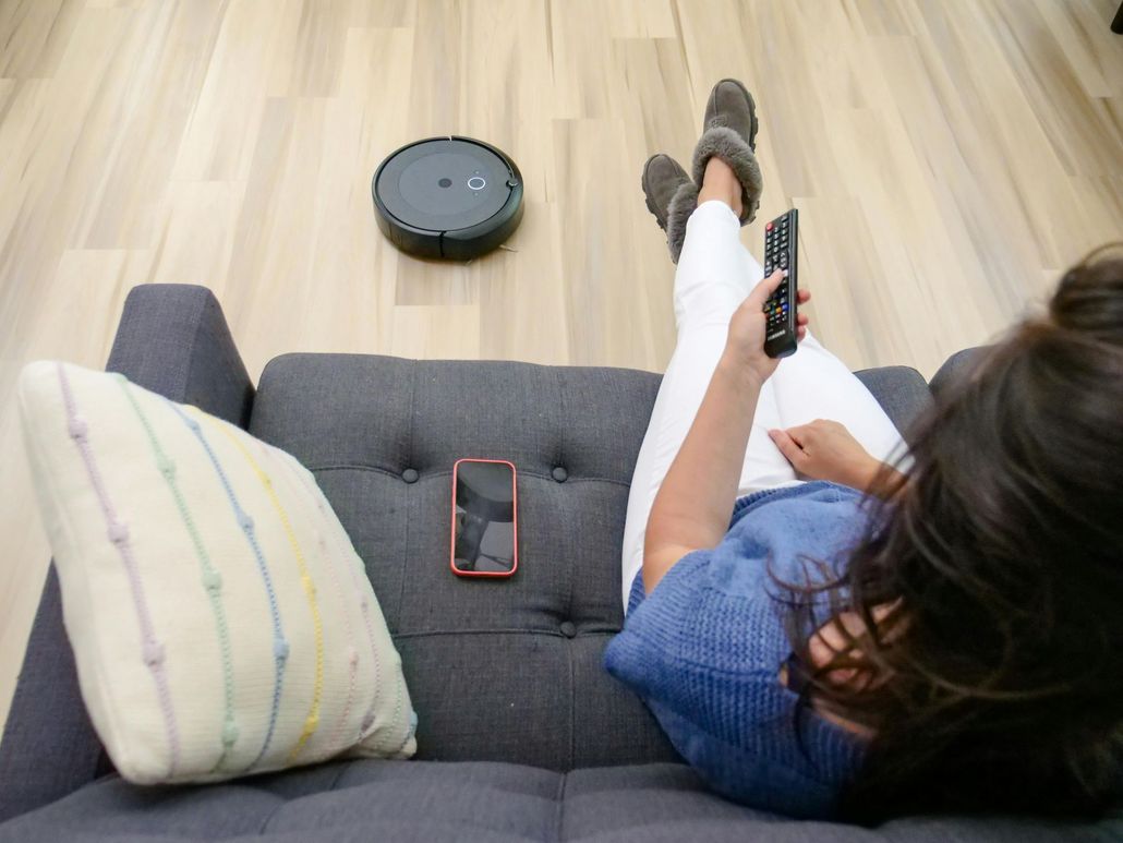 A woman is sitting on a couch holding a remote control while a robot vacuum cleaner is cleaning the floor.