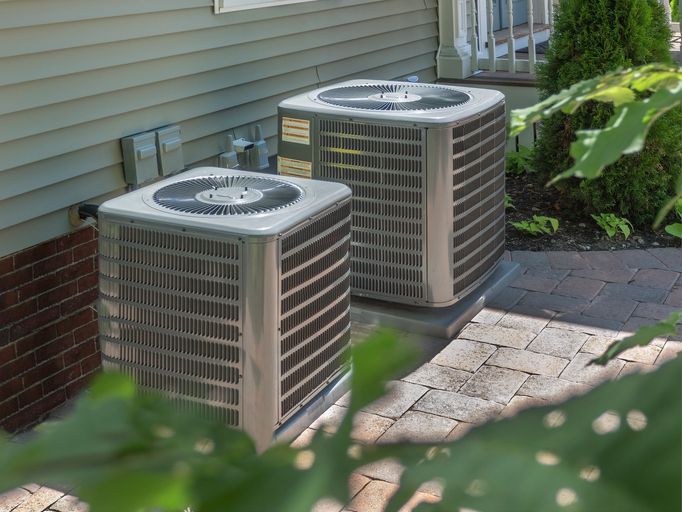 Two outdoor air conditioning units on a brick patio beside a house.