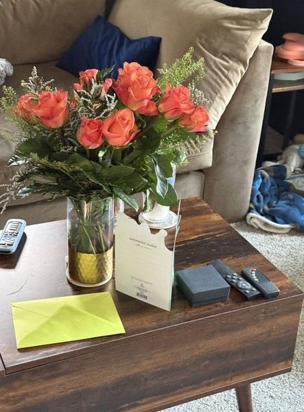 Orange roses in a vase on a coffee table with cards and remotes in a living room.