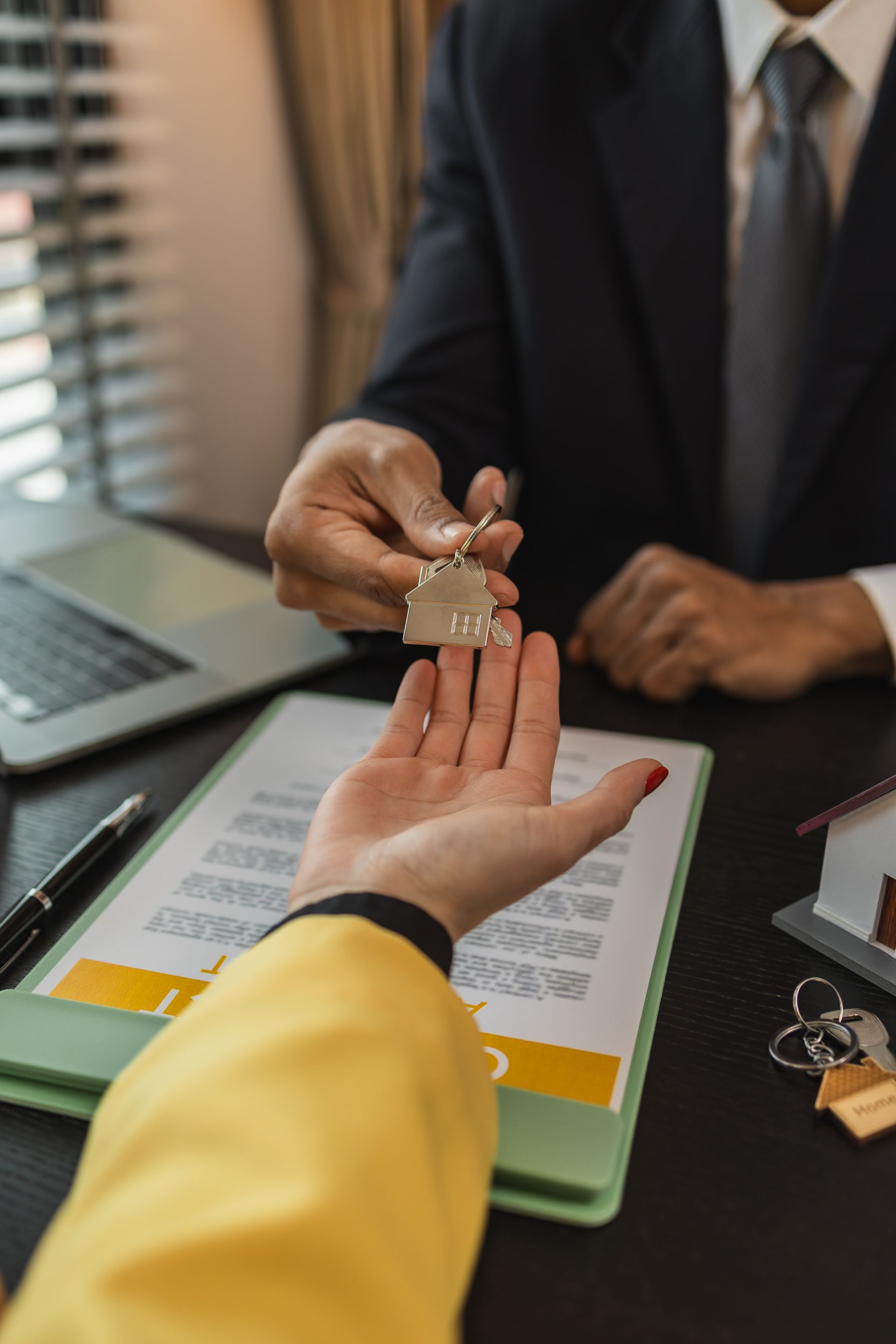 A person in a suit hands house-shaped keys to another person over a desk with a contract and a miniature model home.