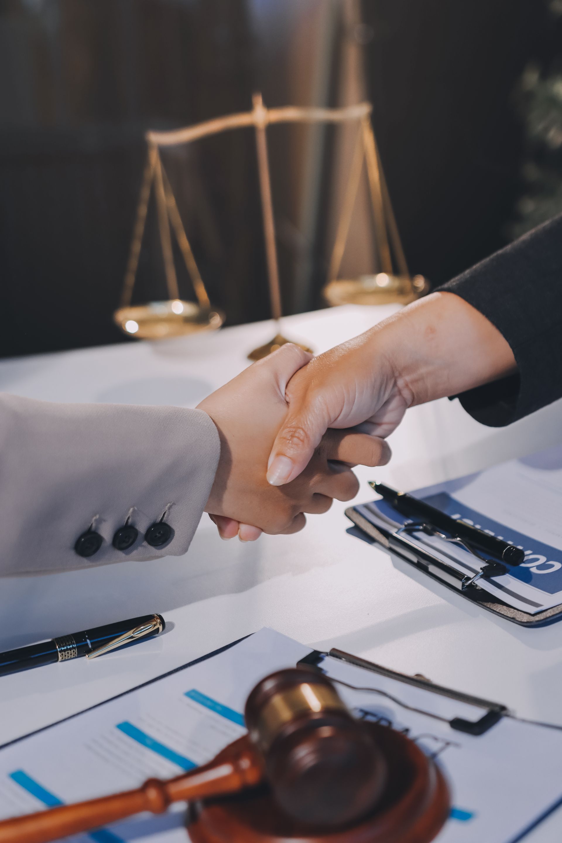 Two professionals shaking hands over a desk with legal documents, a gavel, and a decorative scale of justice. Two professionals shaking hands over a desk with legal documents, a gavel, and a decorative scale of justice.