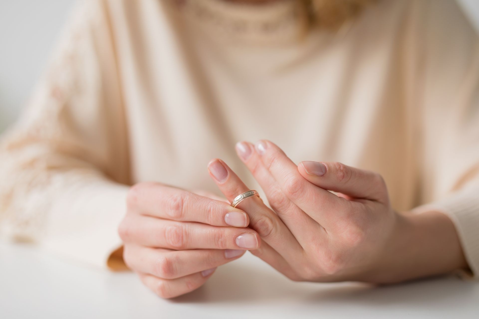 A person wearing a light-colored sweater sits at a white table, gently touching a simple wedding band on their finger. A person wearing a light-colored sweater sits at a white table, gently touching a simple wedding band on their finger.