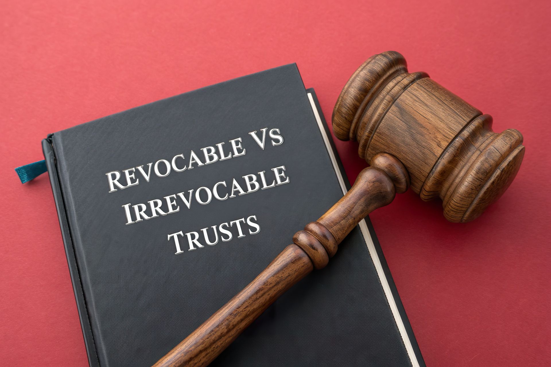 A wooden gavel lies on top of a black book titled Revocable Vs Irrevocable Trusts, set against a red background. A wooden gavel lies on top of a black book titled Revocable Vs Irrevocable Trusts, set against a red background.