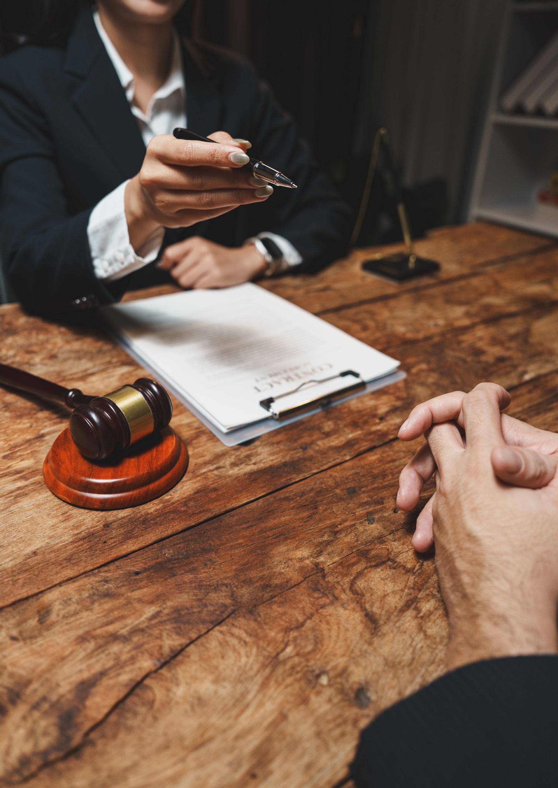 A professional in a suit offers a pen to a person across a wooden table with a contract and a judge's gavel. A professional in a suit offers a pen to a person across a wooden table with a contract and a judge's gavel.