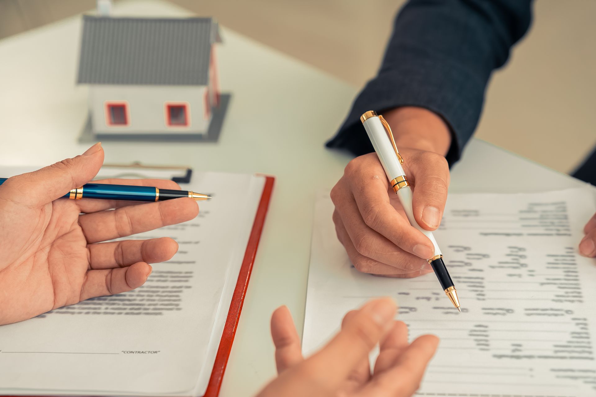 Two people sit at a table signing real estate documents, with a miniature model house in the background.