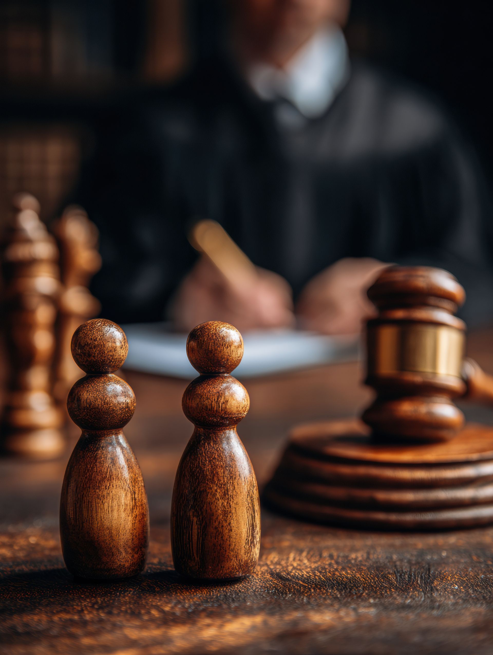 Two wooden chess pawns stand on a table beside a judge's gavel, with a blurred figure writing in the background. Two wooden chess pawns stand on a table beside a judge's gavel, with a blurred figure writing in the background.