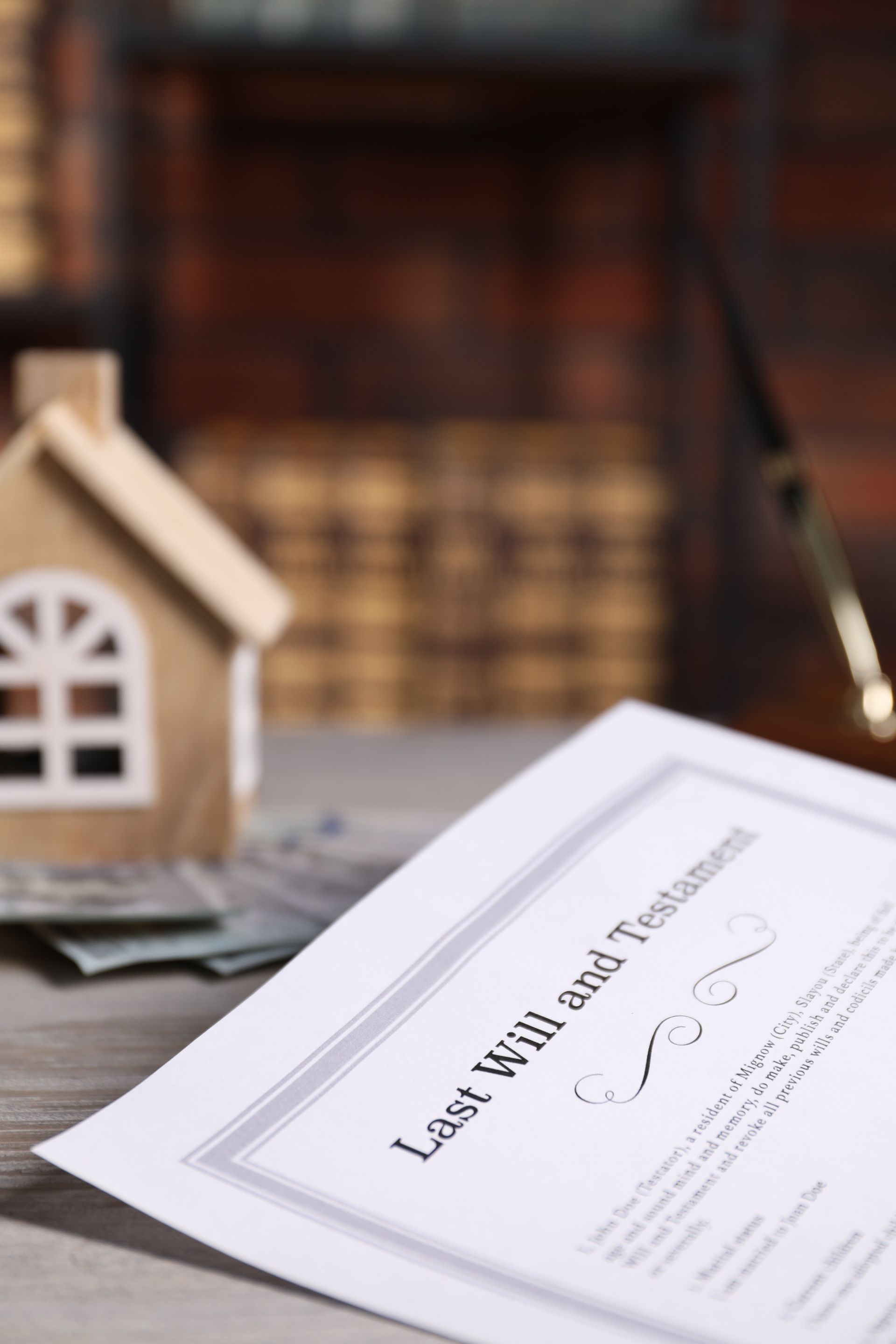 A document labeled Last Will and Testament sits on a table beside a small wooden model house and a stack of money. A document labeled Last Will and Testament sits on a table beside a small wooden model house and a stack of money.
