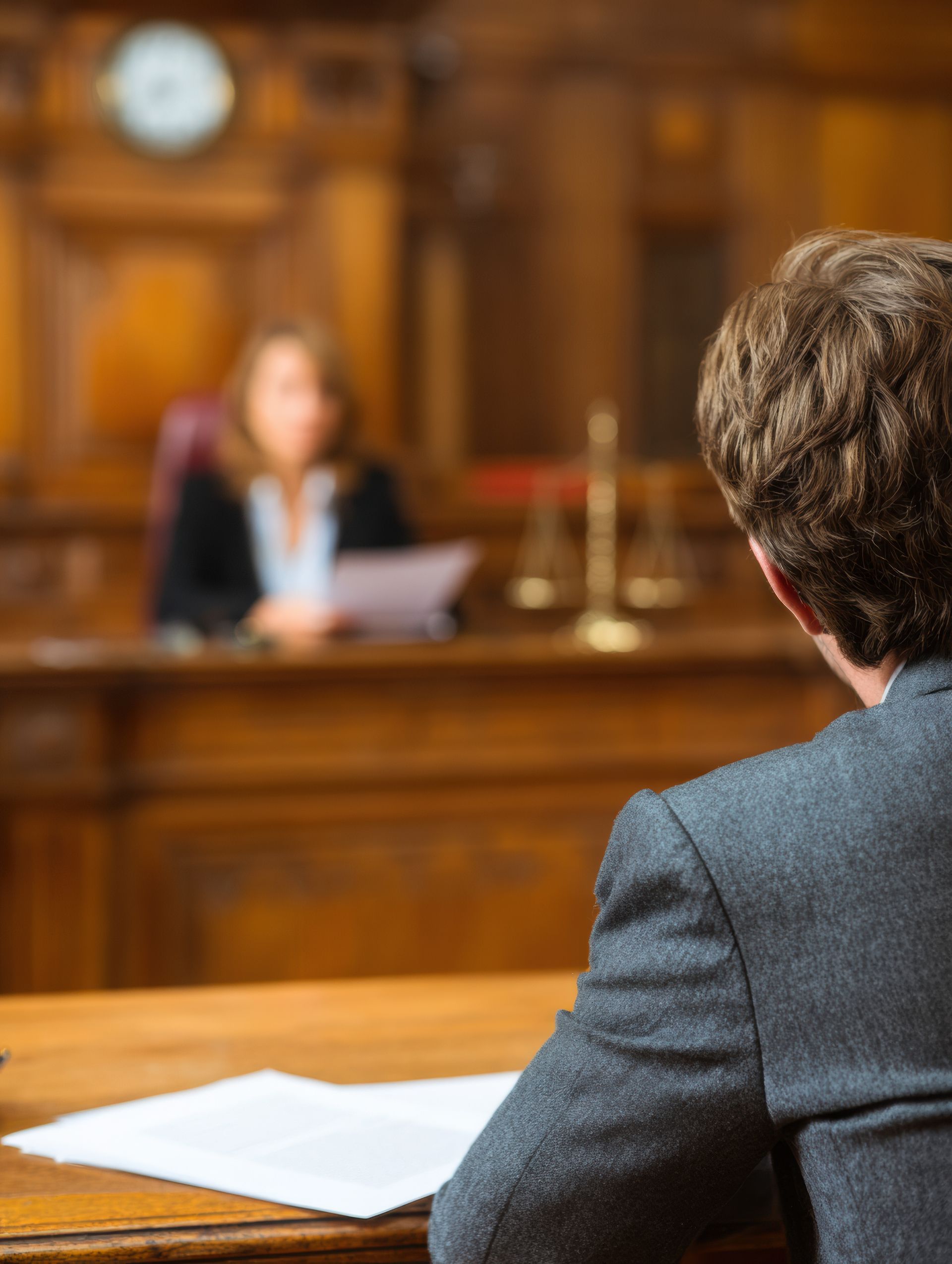 View from behind a person in a suit, sitting at a desk with papers, facing a judge in a wood-paneled courtroom. A golden balance scale rests on a thick book next to a wooden gavel against a solid sage green background.