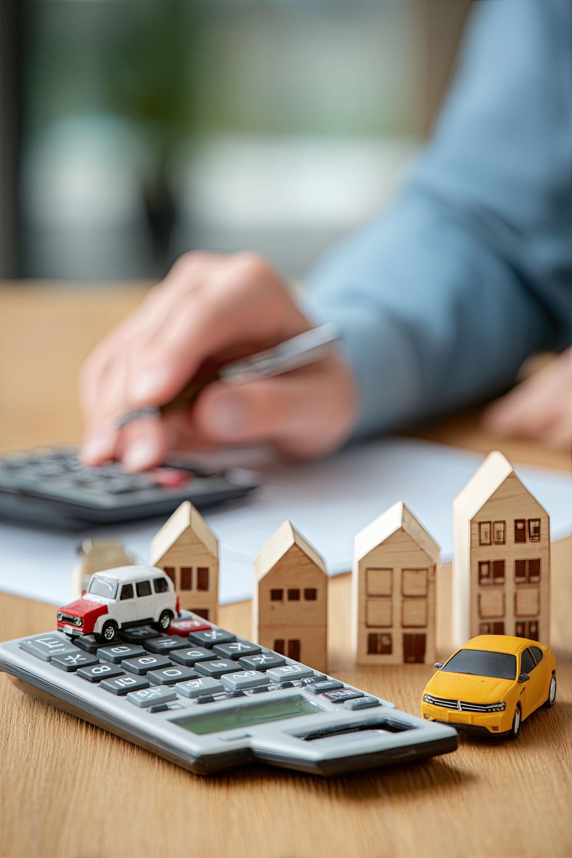 A person uses a calculator and pen next to miniature wooden house models and toy cars on a wooden table. A person uses a calculator and pen next to miniature wooden house models and toy cars on a wooden table.