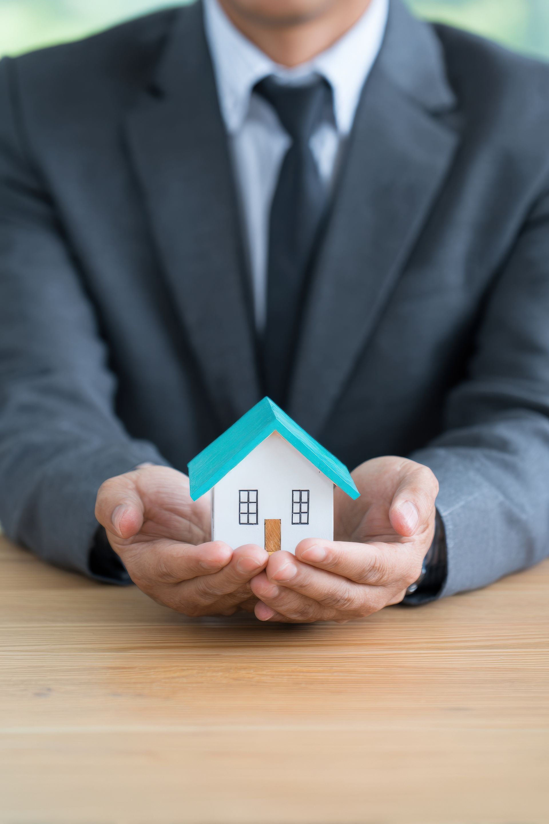 A person in a business suit holds a small, white model house with a teal roof in their cupped hands.