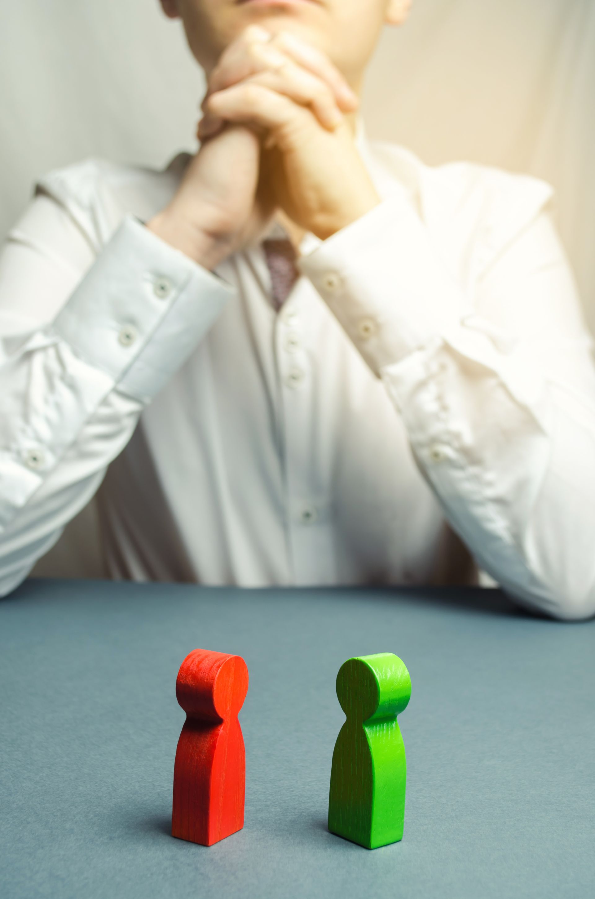 A person sits with hands clasped behind a red and a green wooden figure placed on a table. A person sits with hands clasped behind a red and a green wooden figure placed on a table.