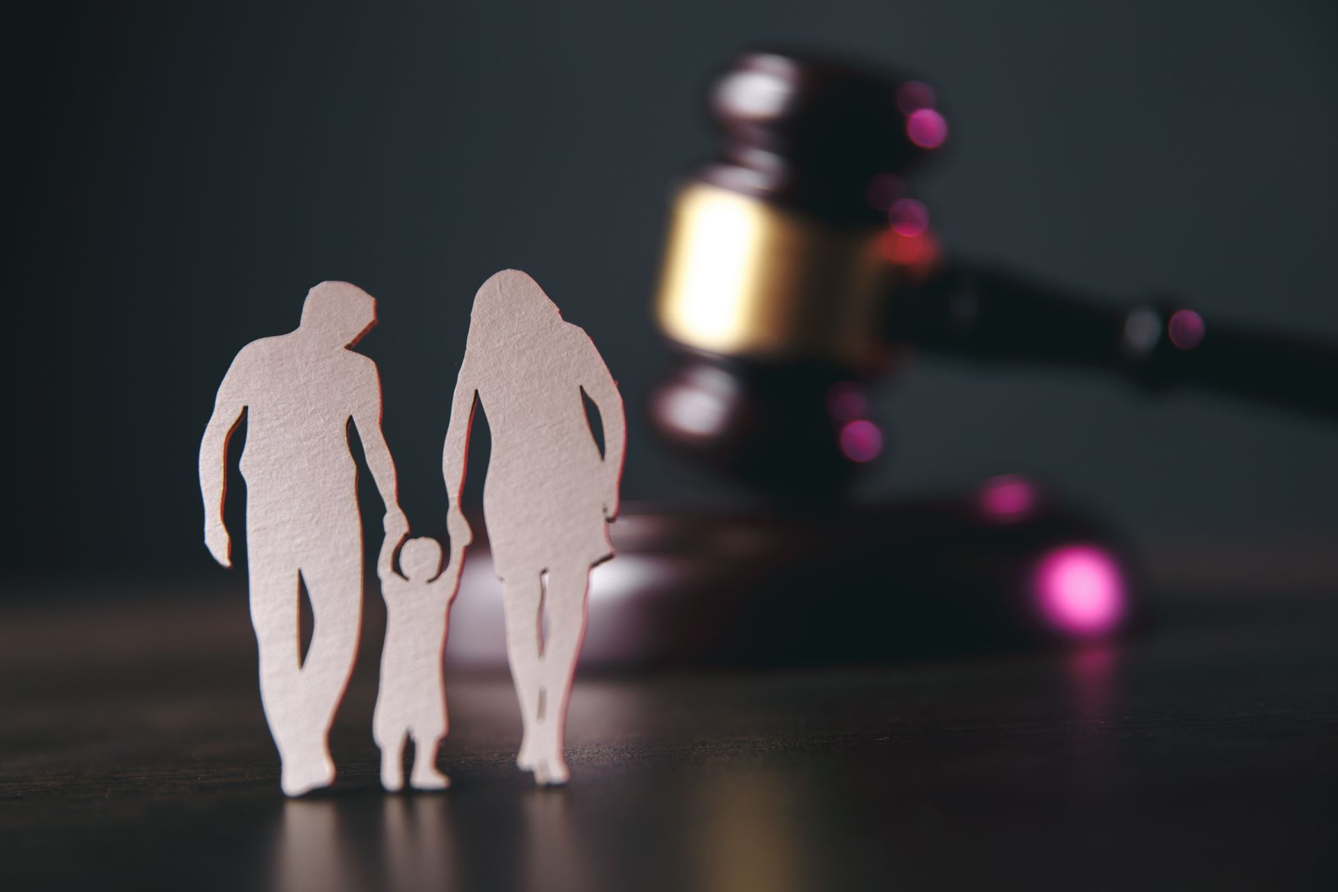 Paper cutouts of two adults and a child standing in front of a blurred wooden judge's gavel on a dark surface.