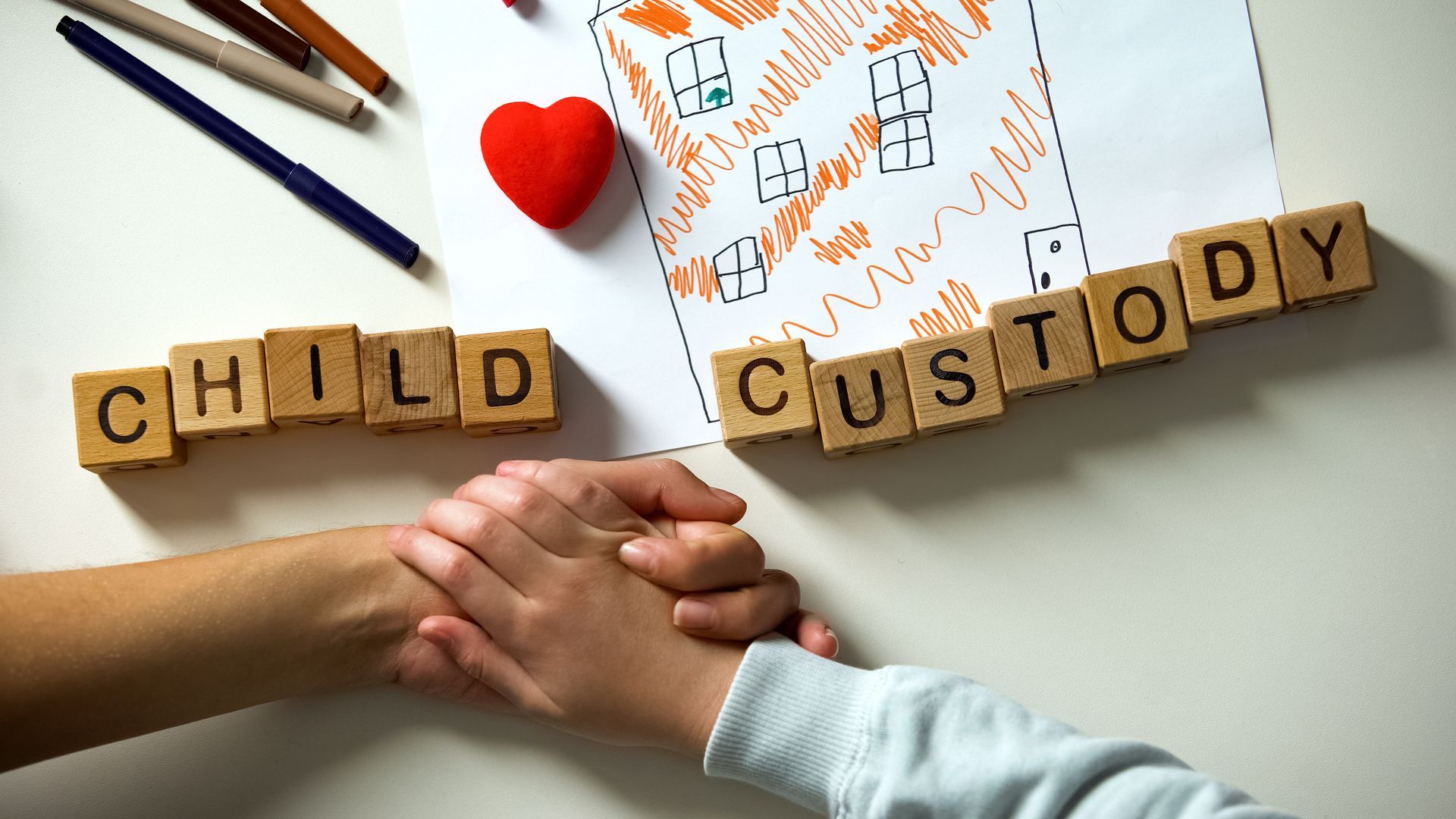 Two hands held together over wooden blocks spelling CHILD CUSTODY, next to a child's drawing of a house and a red heart.