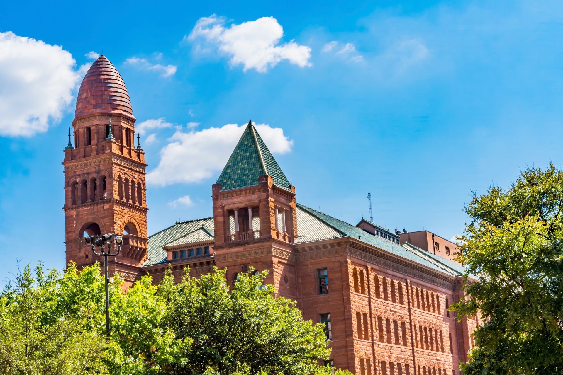 A historic red brick building with a tall, domed tower and green shingled roofs under a bright blue sky with clouds.