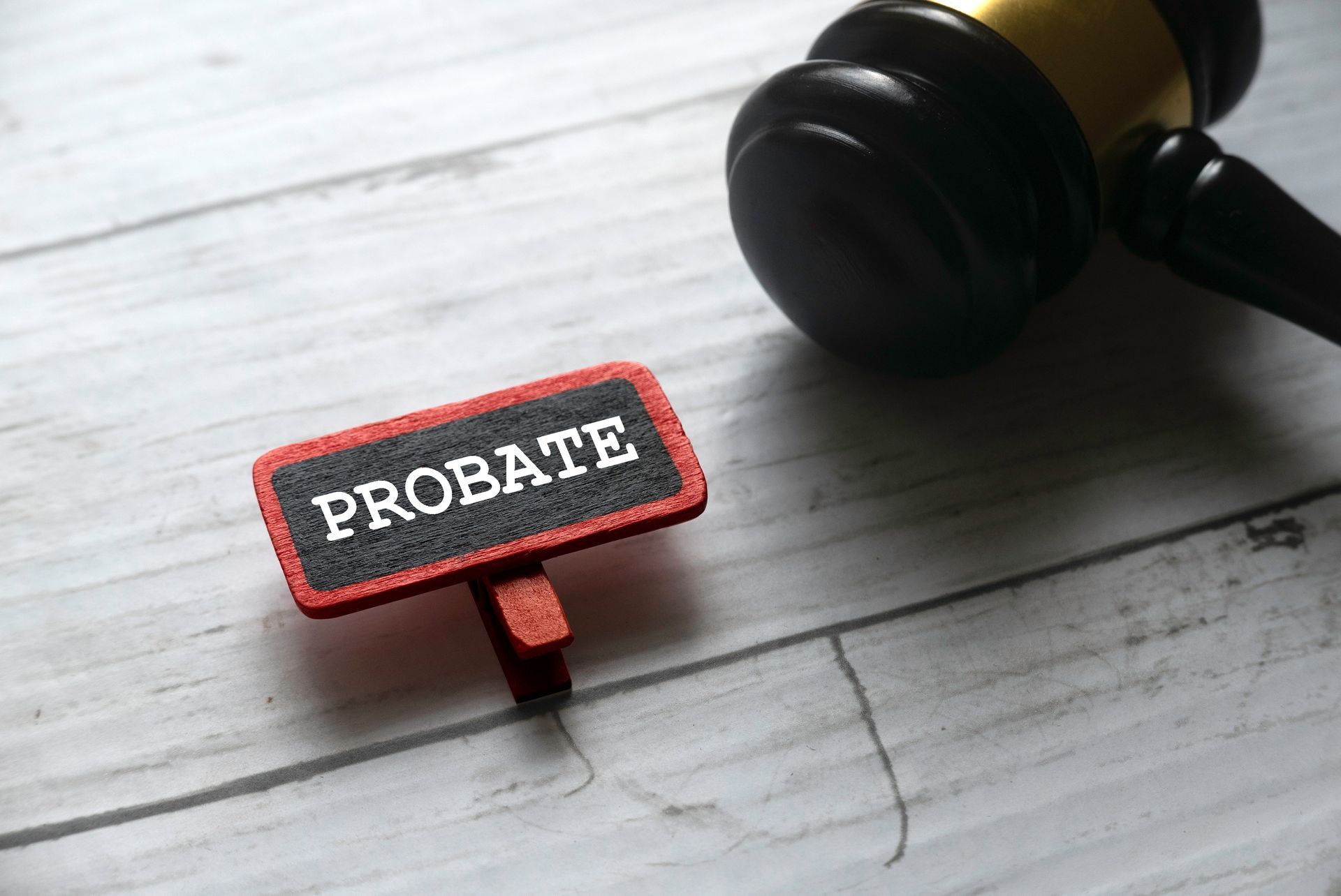 A wooden gavel lies beside a small red-bordered chalkboard sign that says PROBATE on a light wood grain surface.
