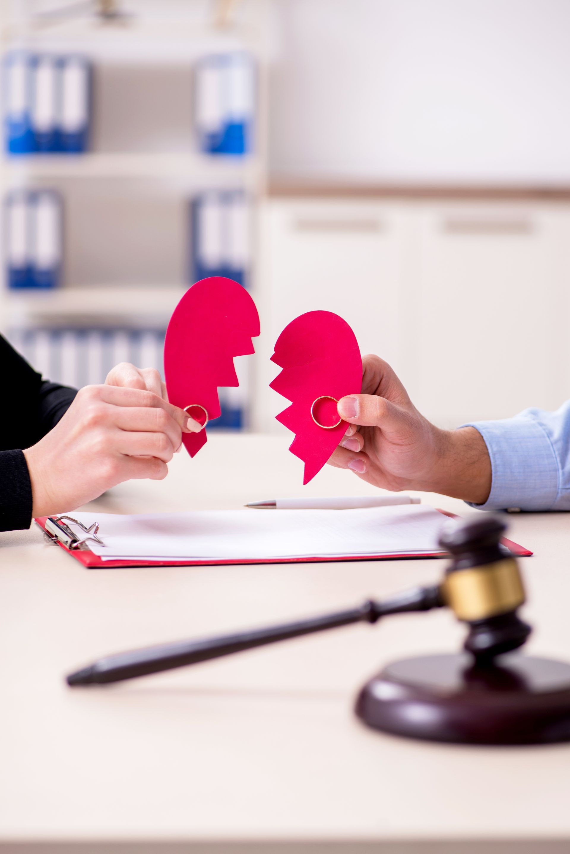 Two people in a legal setting hold halves of a broken red paper heart with wedding rings, near a judge’s gavel. Two people in a legal setting hold halves of a broken red paper heart with wedding rings, near a judge’s gavel.