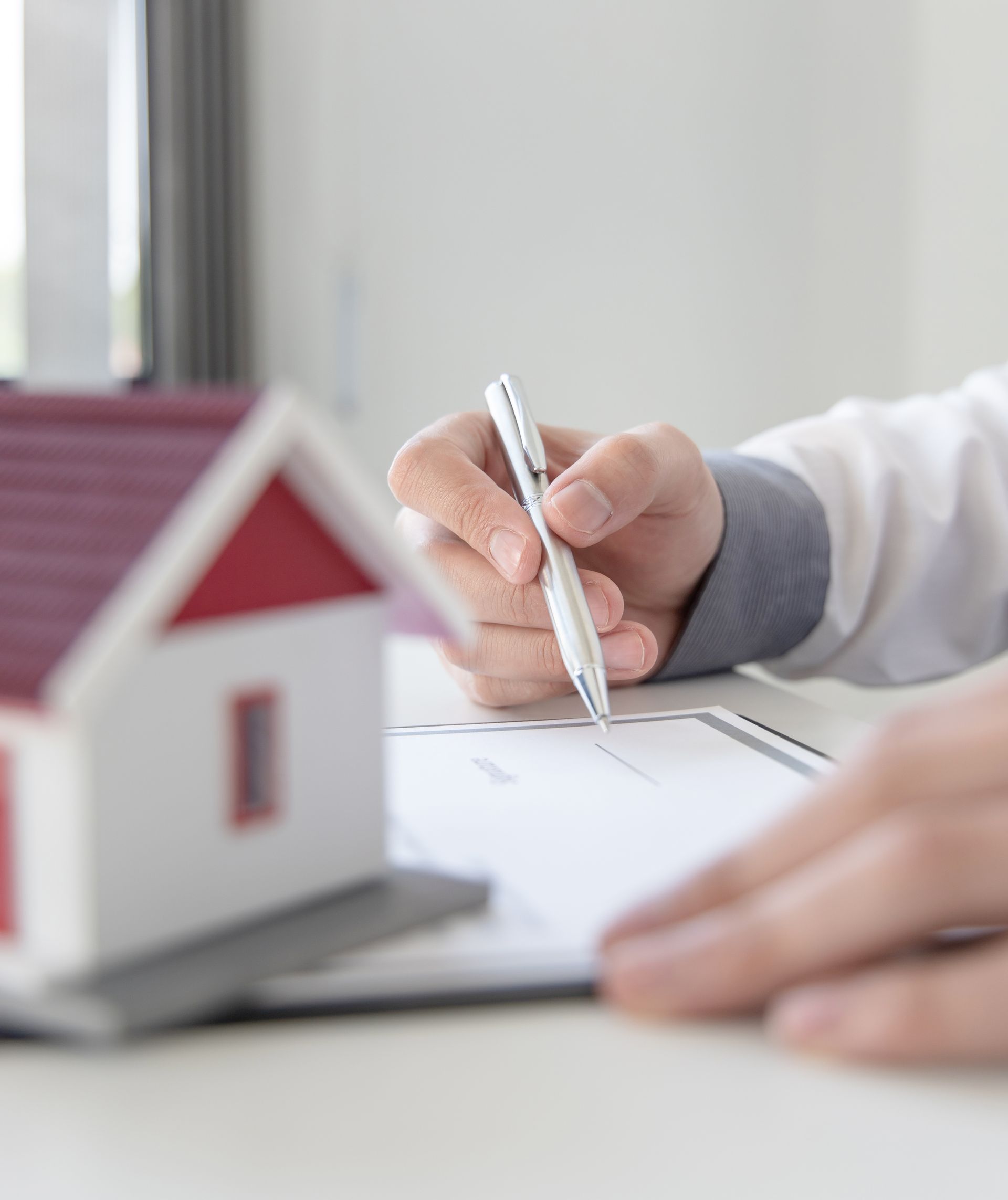 A person in a white sleeve signs a document next to a small model house with a red roof on a white surface.