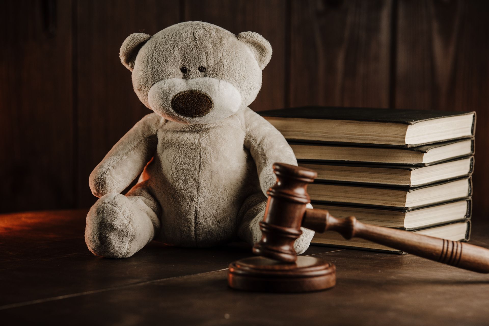 A teddy bear sits on a wooden surface next to a gavel and a small stack of books against a dark, moody background.