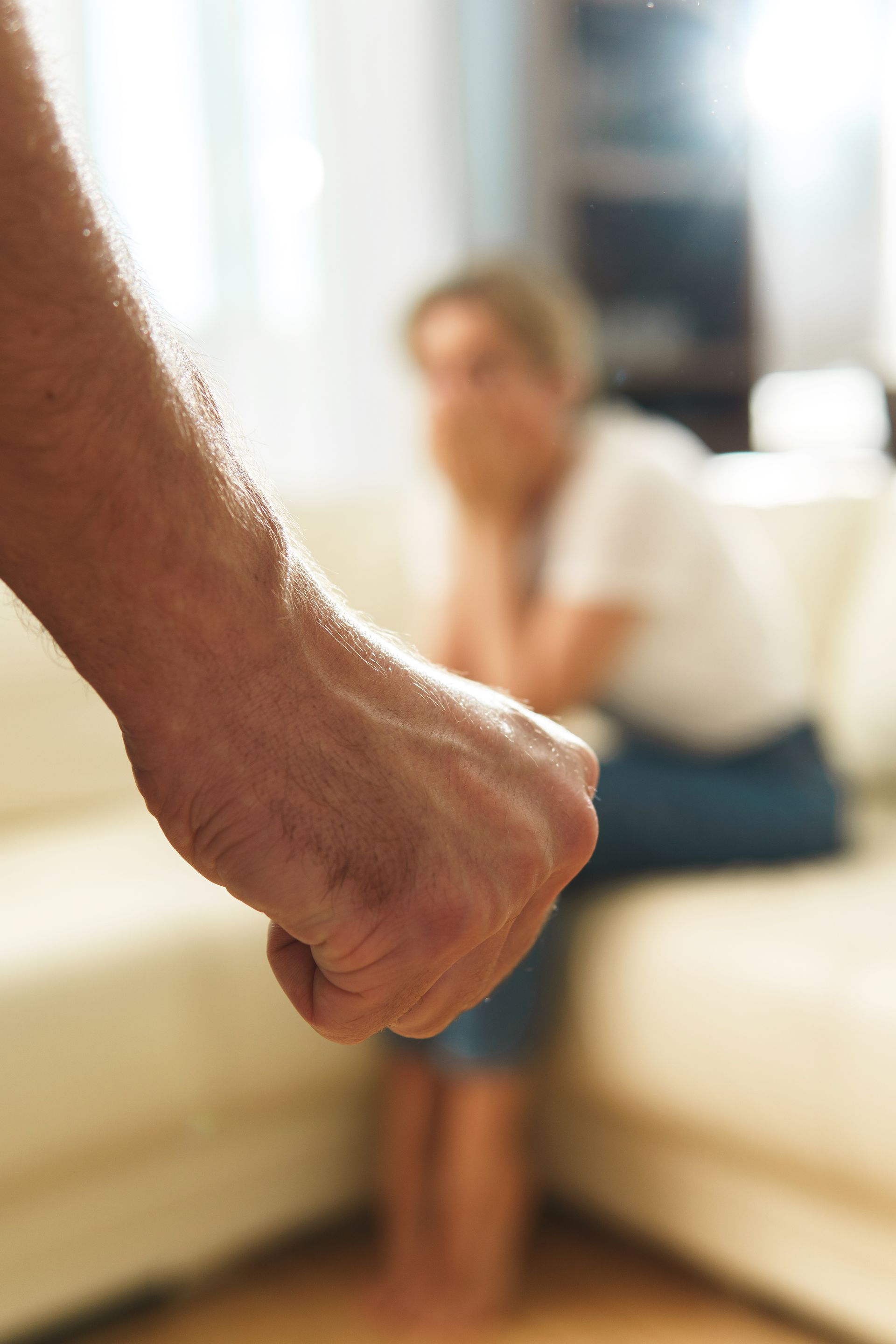 A clenched fist in the foreground with an out-of-focus person sitting in the background. A clenched fist in the foreground with an out-of-focus person sitting in the background.