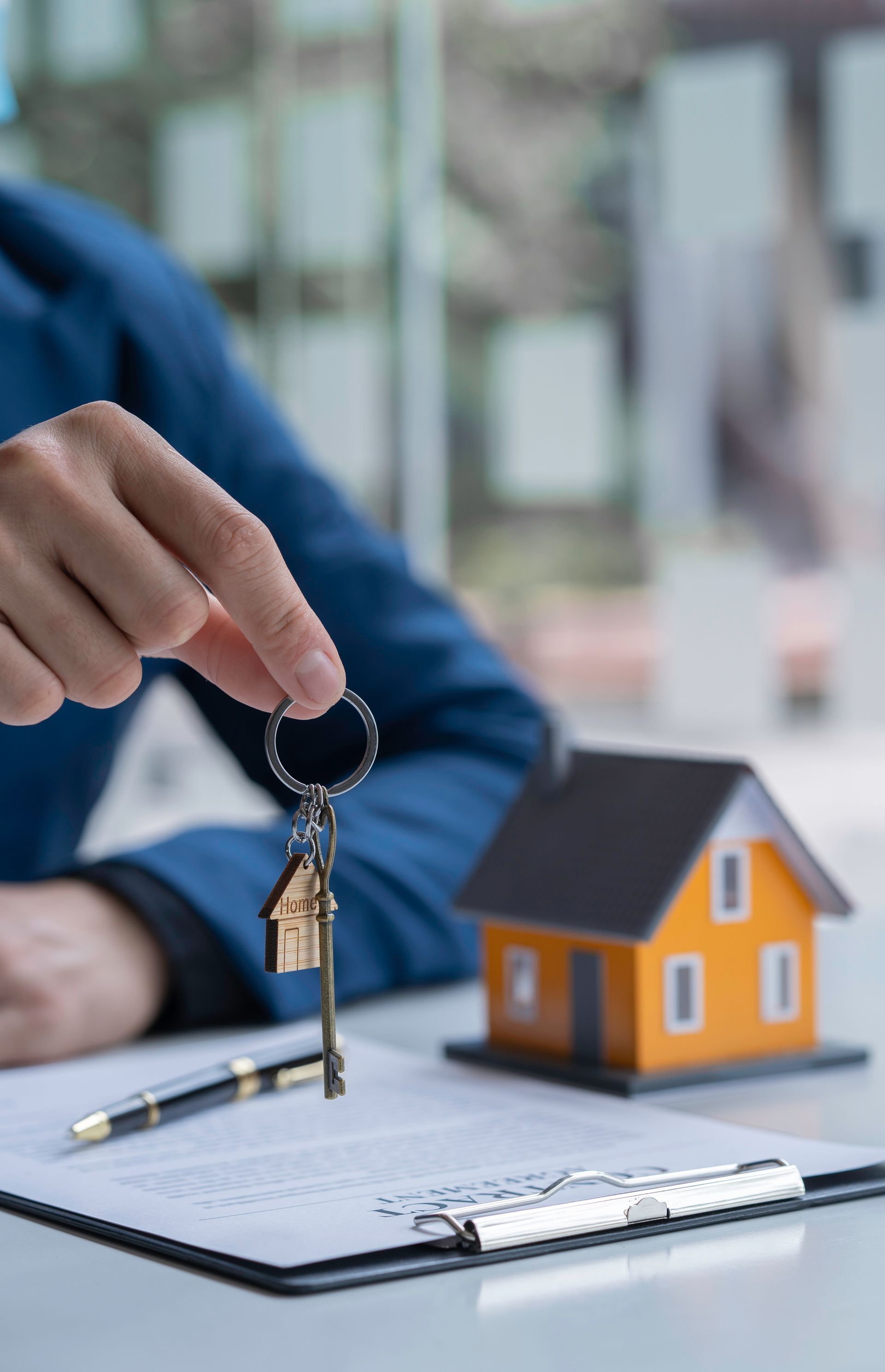 A person in a blue jacket holds house-shaped keys over a document, next to a small orange house model on a desk.