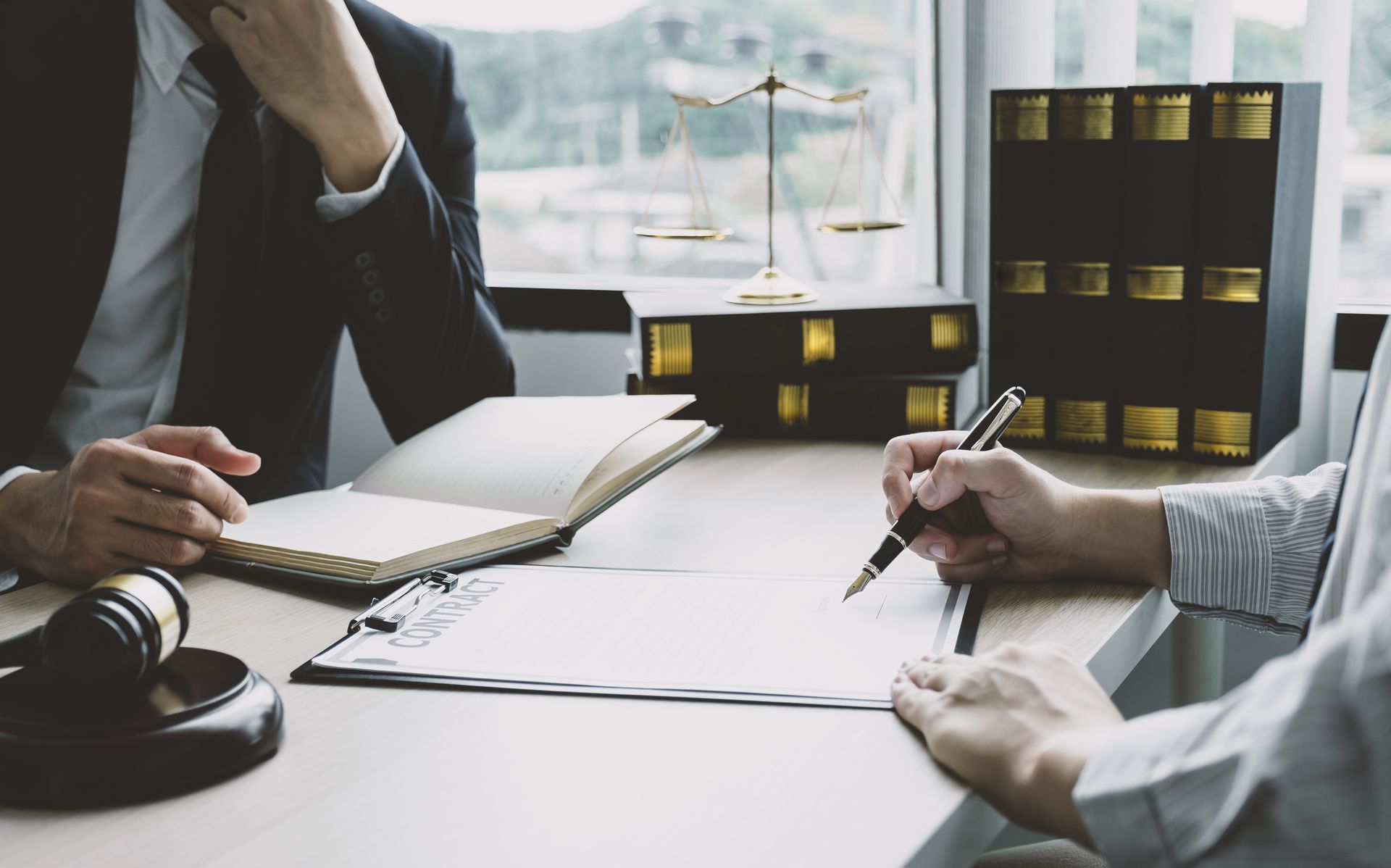 A lawyer and client sit at a desk with law books, a gavel, and a balance scale while the client signs a document. A lawyer and client sit at a desk with law books, a gavel, and a balance scale while the client signs a document.