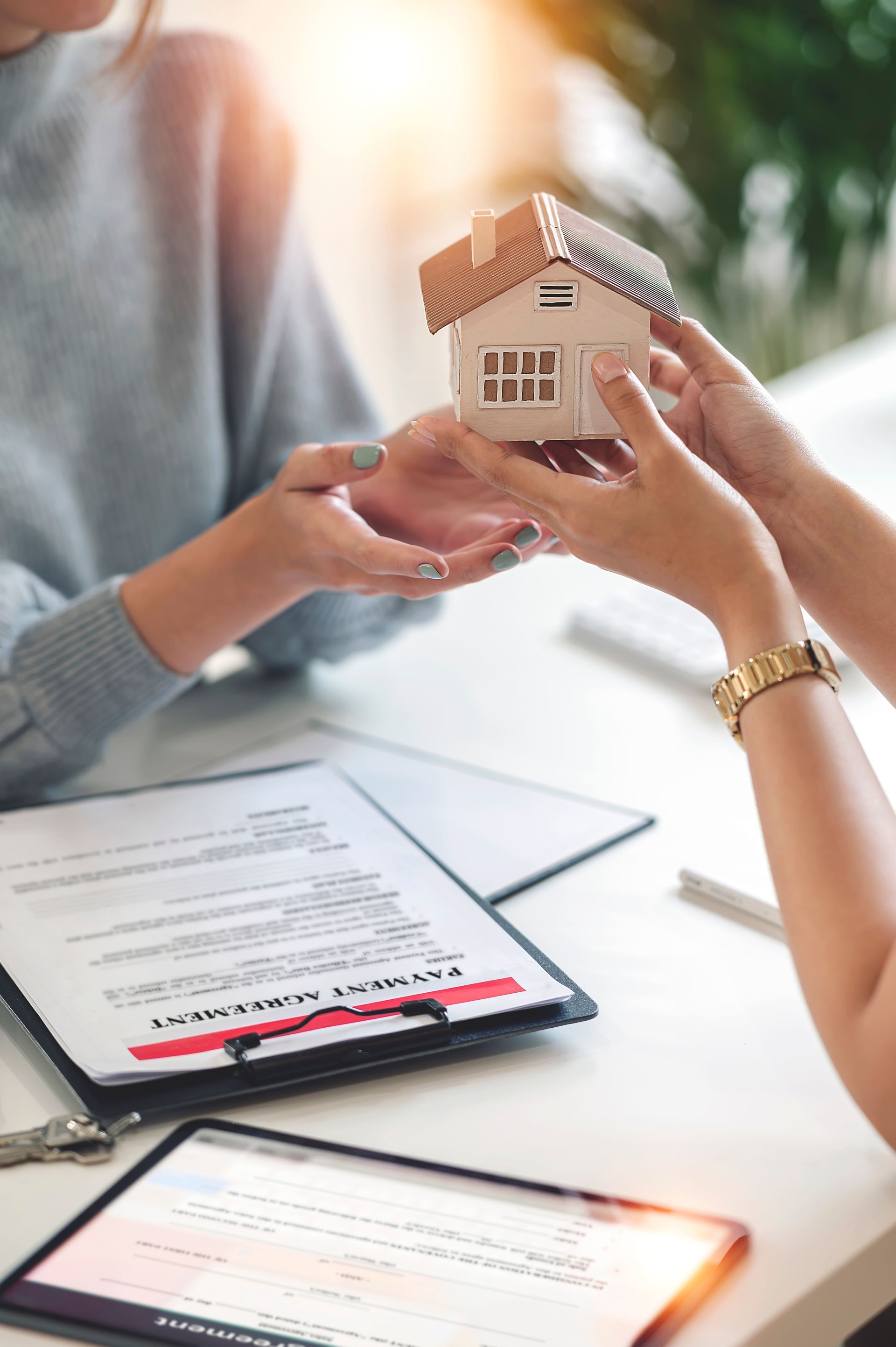 Two people exchange a small model house over a table with documents and a tablet, signifying a real estate transaction.