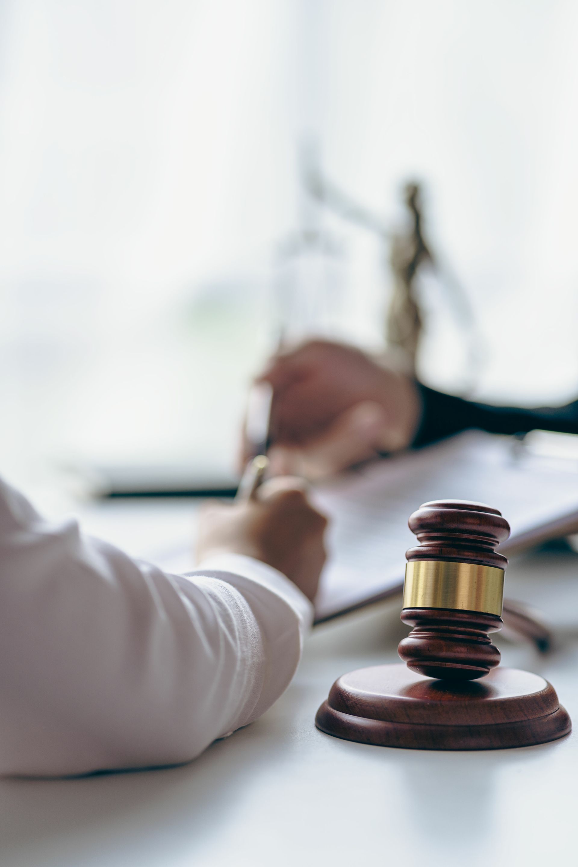 A judge's gavel rests on a white desk in the foreground, with blurred figures signing legal documents in the background. A judge's gavel rests on a white desk in the foreground, with blurred figures signing legal documents in the background.