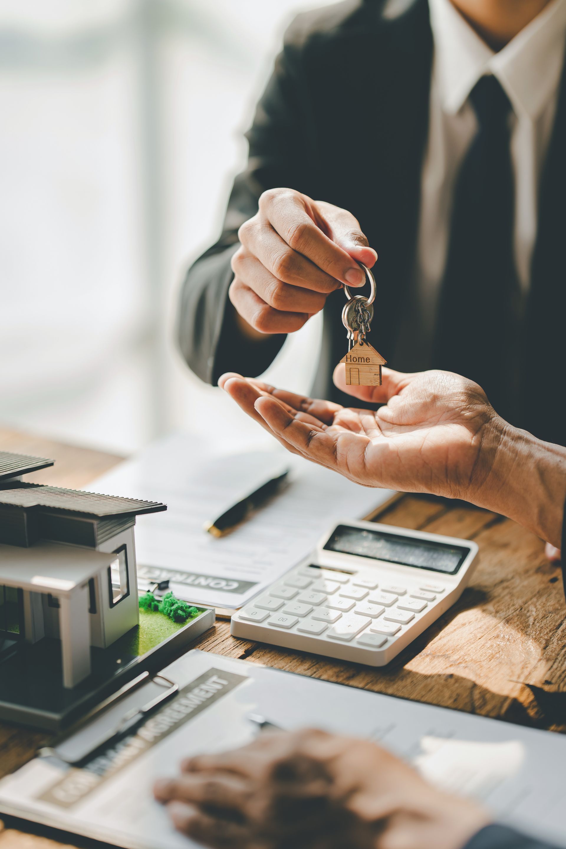 A professional in a suit hands house keys to another person over a desk with a house model, calculator, and documents.