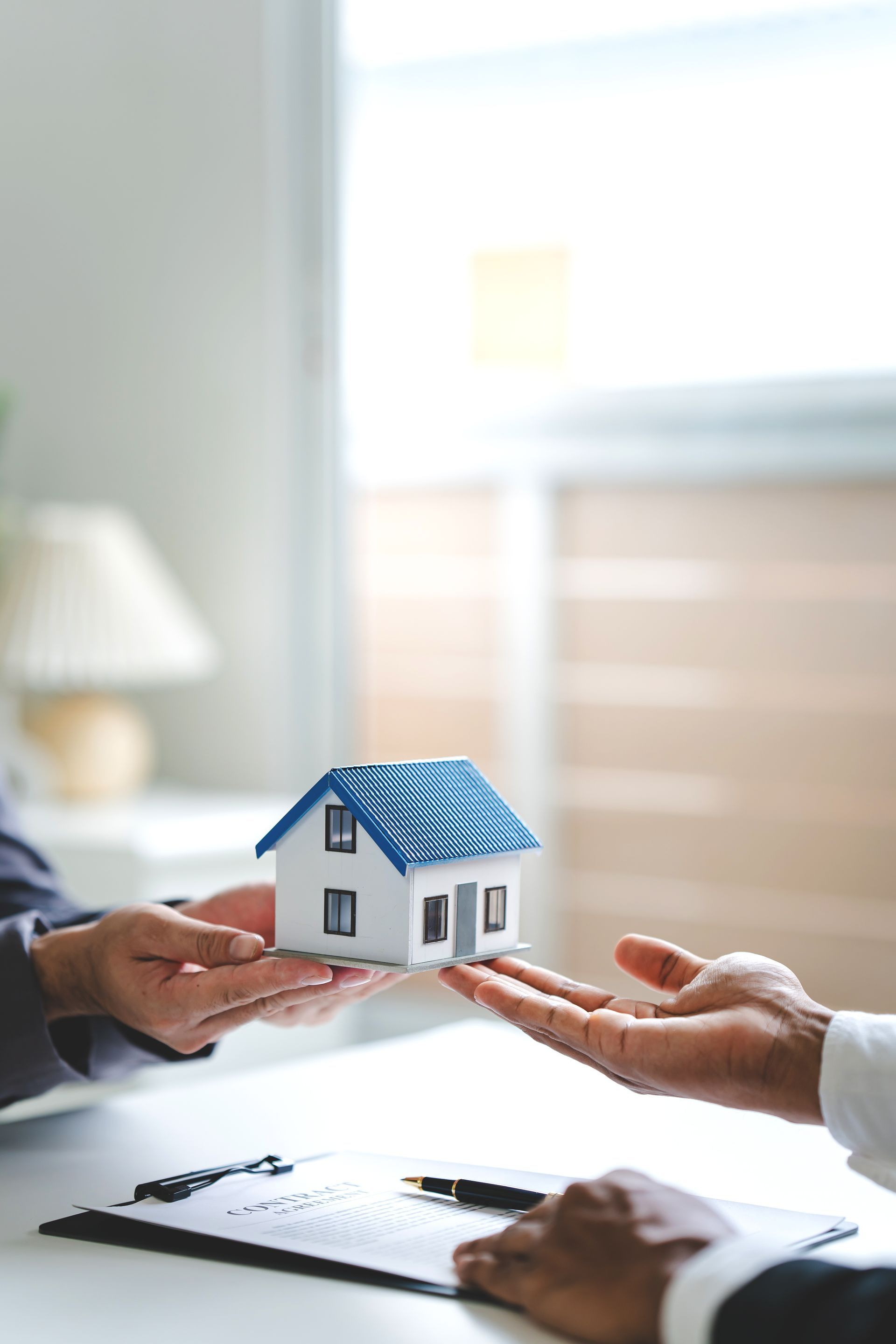 Two people exchange a small model house over a desk with paperwork, representing a real estate or insurance transaction.