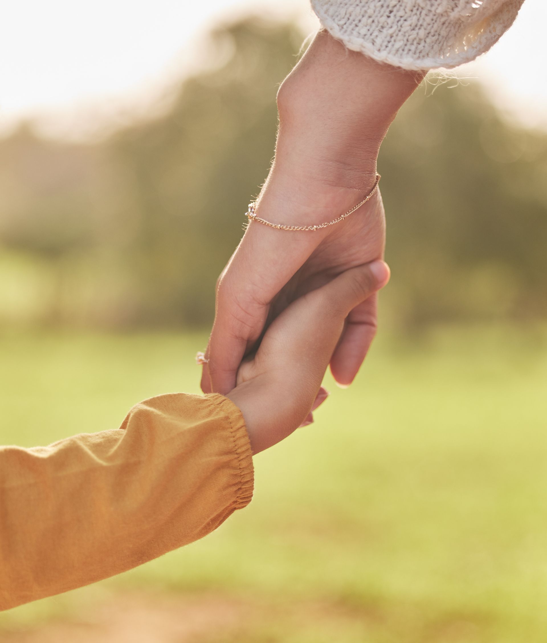 An adult wearing a light sweater and bracelet holds the hand of a person wearing a yellow long-sleeved shirt outdoors.
