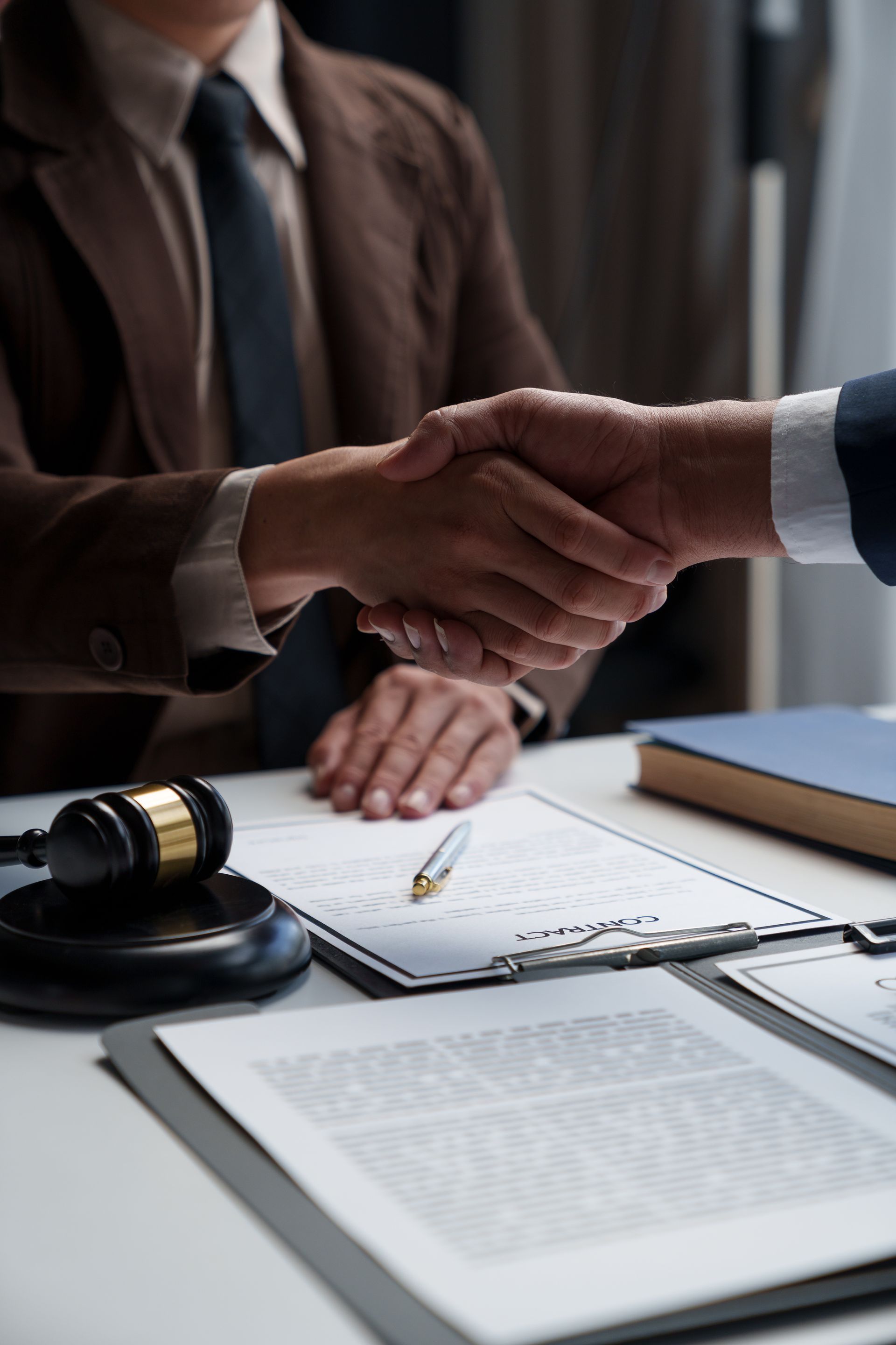 Two professionals in suits shake hands over a desk with legal documents and a gavel, symbolizing a finalized agreement.