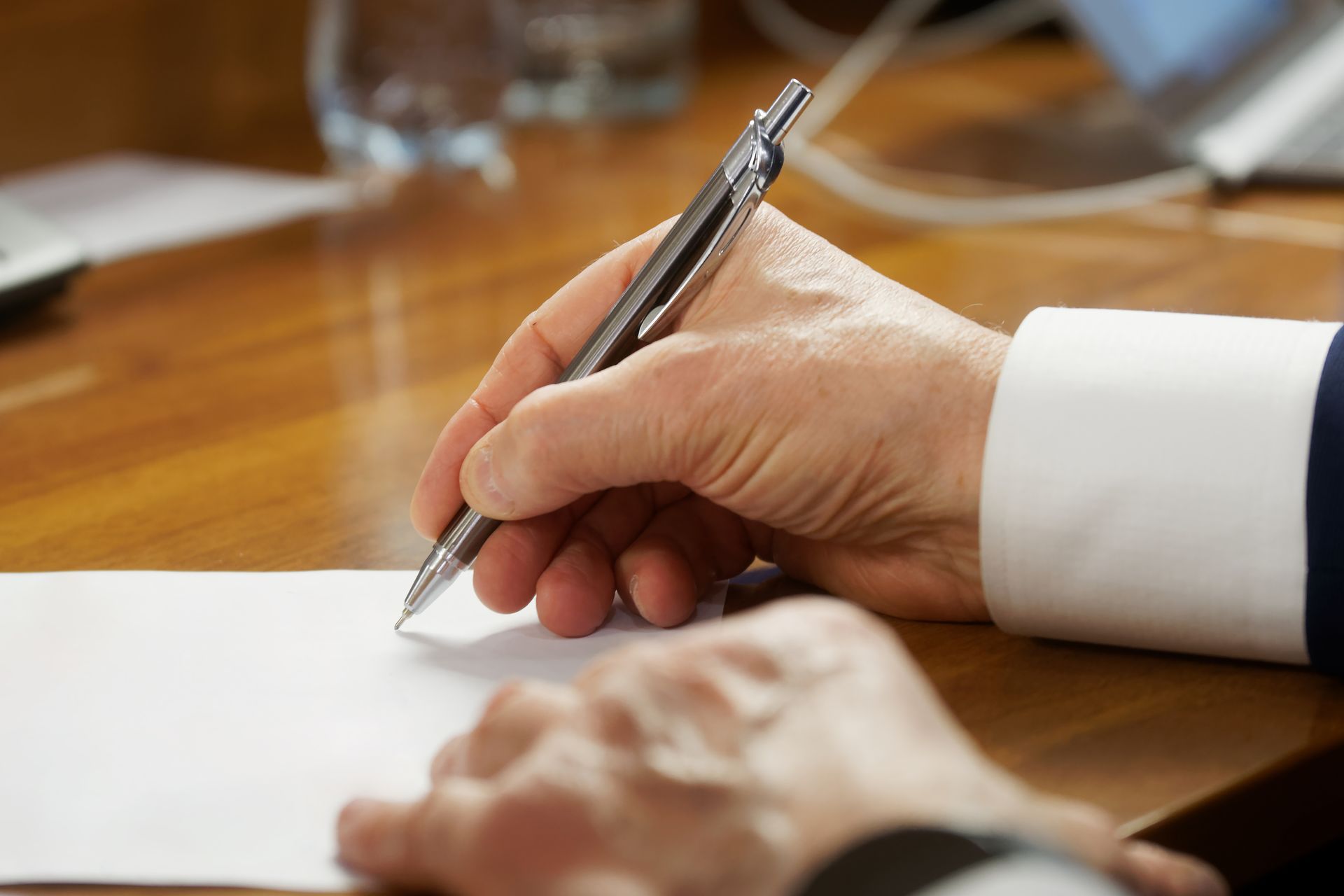 A close-up of a person's hand wearing a white cuff writing on a white sheet of paper with a metallic pen at a desk. A close-up of a person's hand wearing a white cuff writing on a white sheet of paper with a metallic pen at a desk.