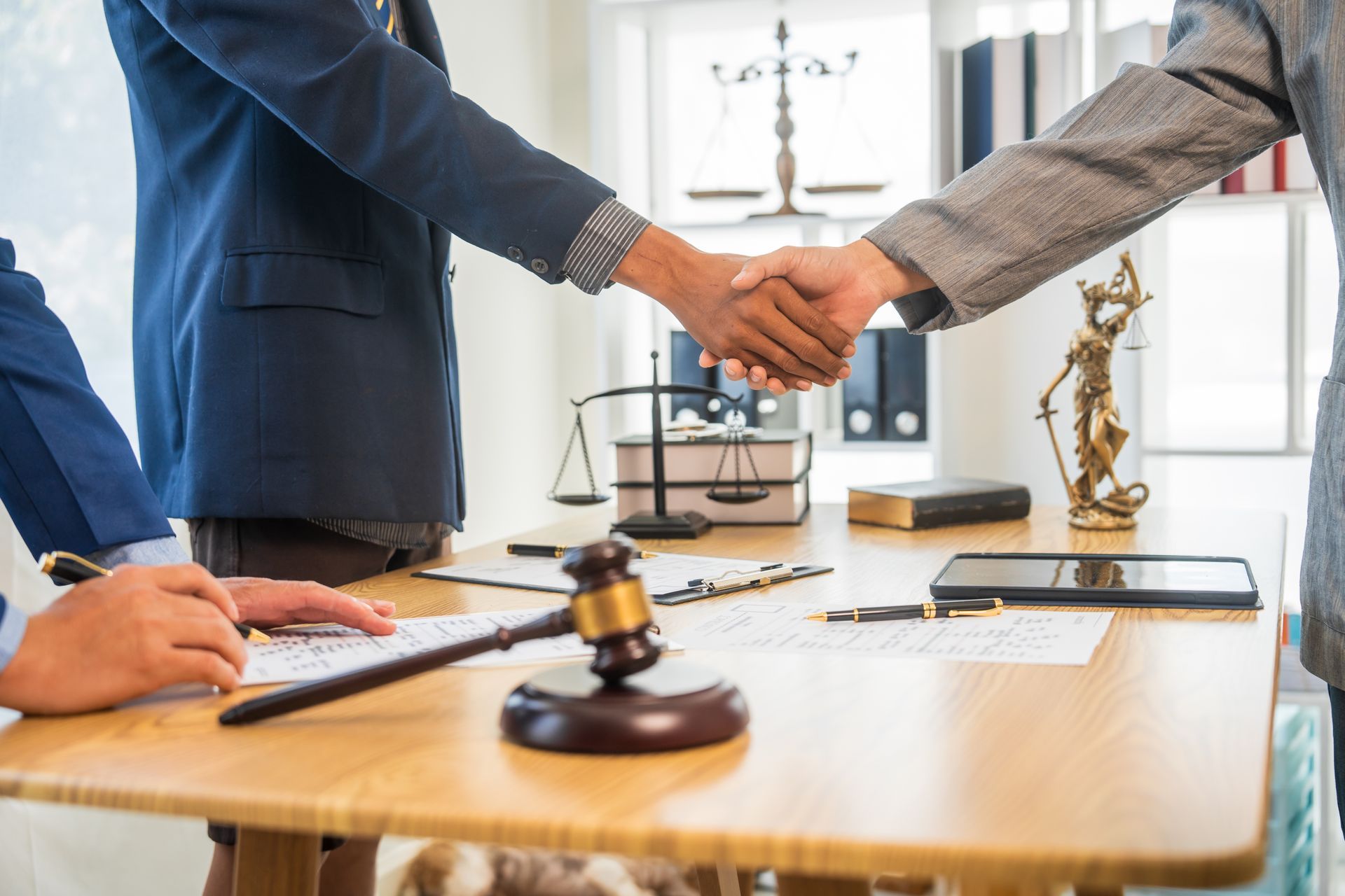 Two people in business attire shake hands over a wooden desk with a gavel, legal scales, and a statue of Lady Justice.