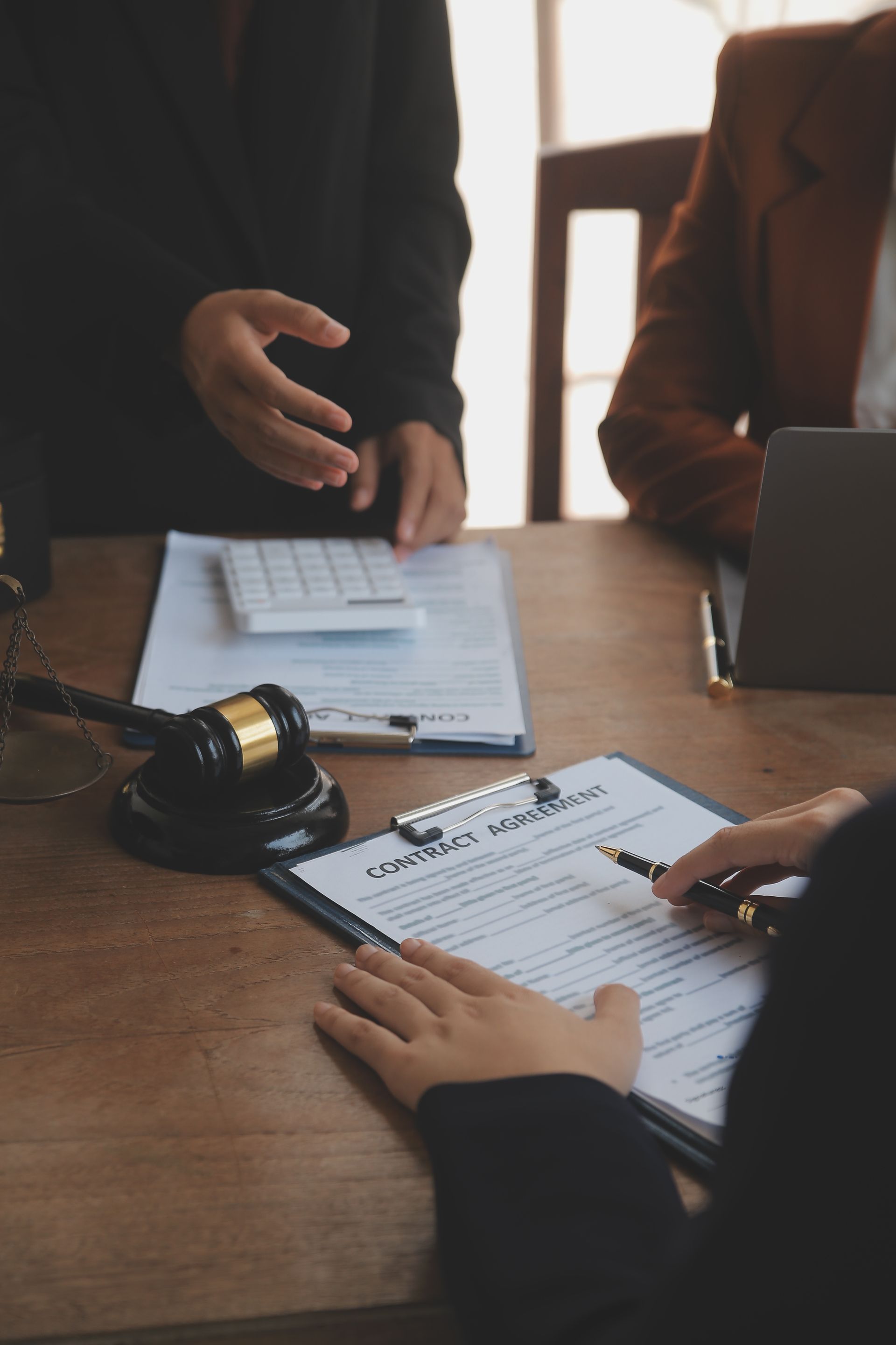 Two professionals sit at a desk with a gavel and document, one signing paperwork while the other gestures during a meeting.