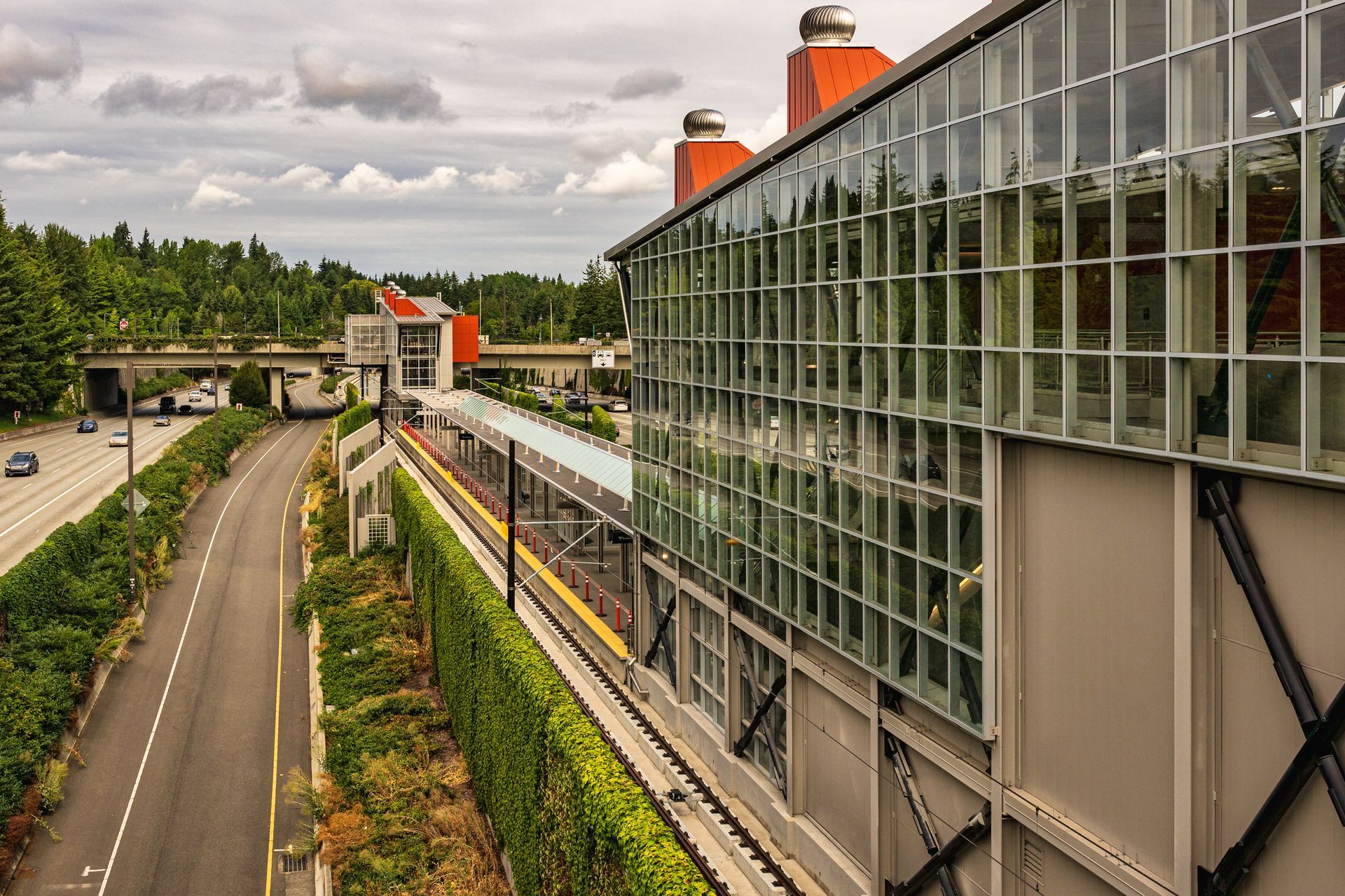 A modern transit station with a large glass facade sits beside a highway and a green, tree-lined landscape under a sky.
