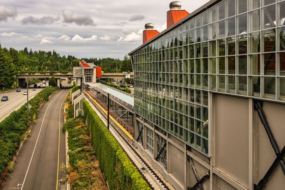 A modern transit station with a large glass facade sits beside a highway and a green, tree-lined landscape under a sky. A modern transit station with a large glass facade sits beside a highway and a green, tree-lined landscape under a sky.