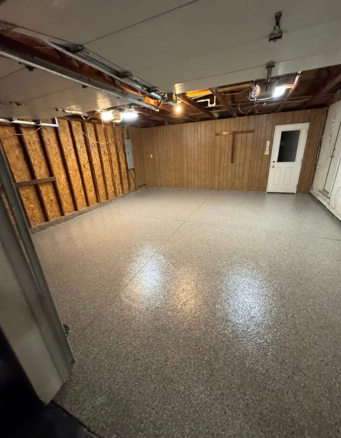 Empty garage with gray speckled floor, exposed ceiling, wood-paneled wall, and a white door.