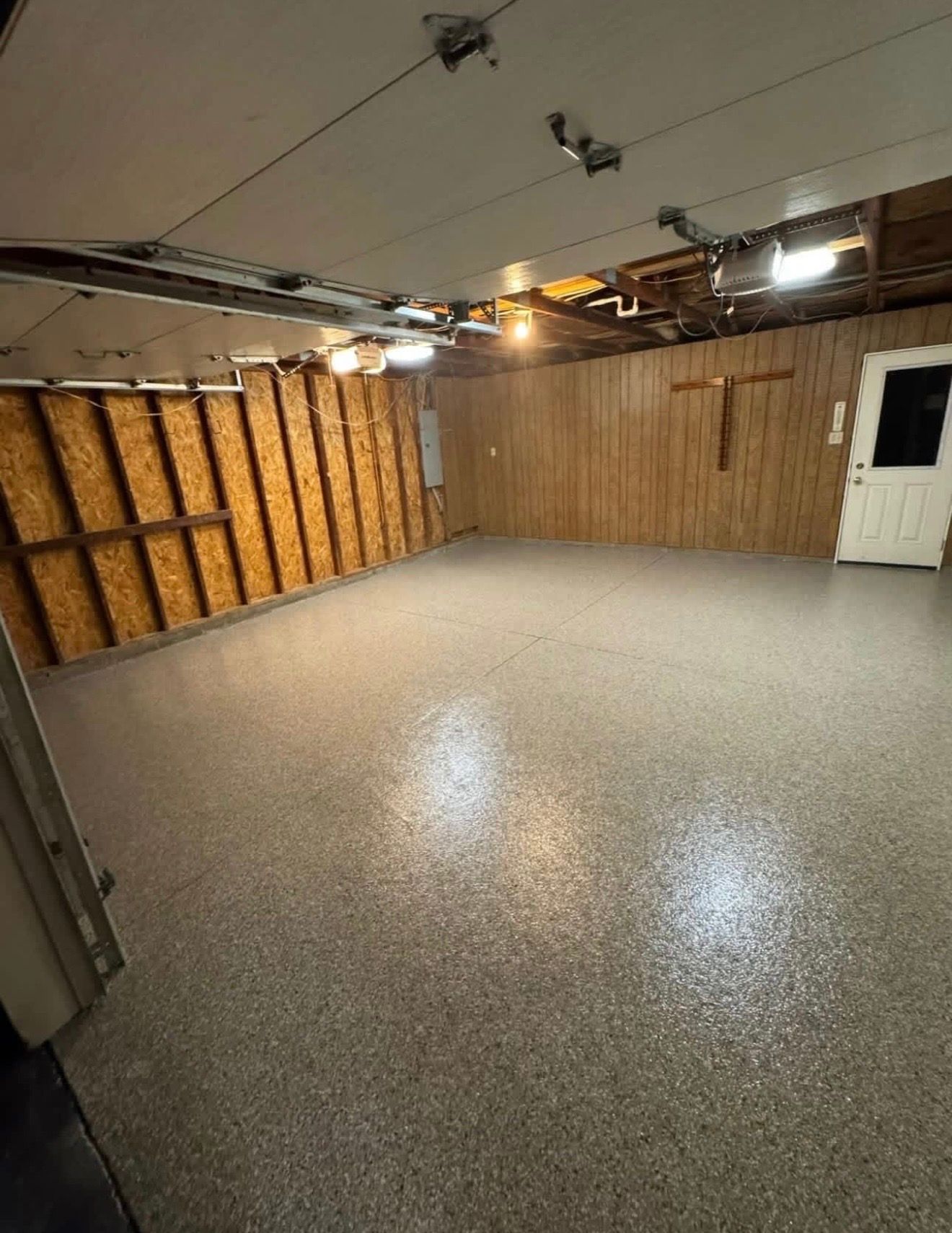 Empty garage with speckled concrete floor, exposed ceiling, wood-paneled walls, and a white side door.