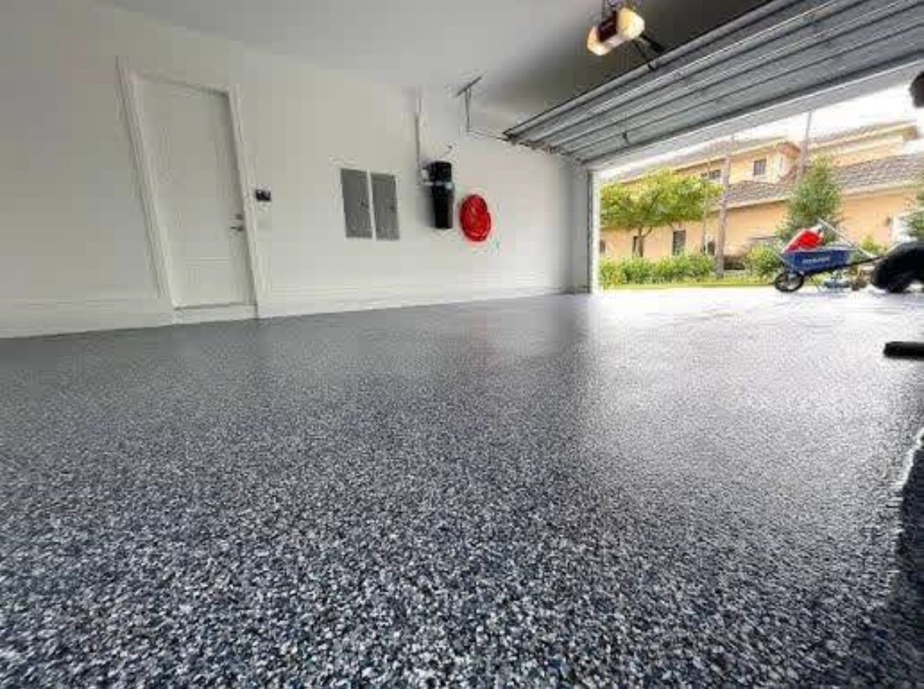 Empty garage with speckled epoxy floor, white walls, and open door to a driveway and house outside