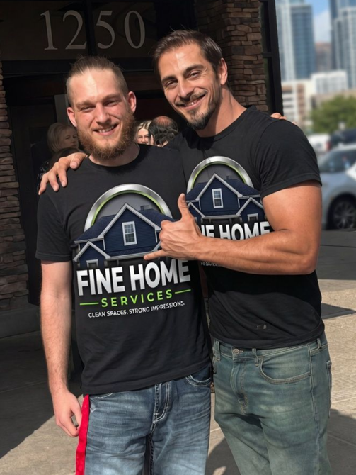 Two men in black Fine Home Services shirts smiling and posing outdoors in front of a building.
