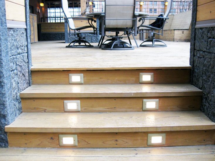 A set of three wooden stairs featuring built-in, glowing recessed lights on a patio with outdoor furniture.