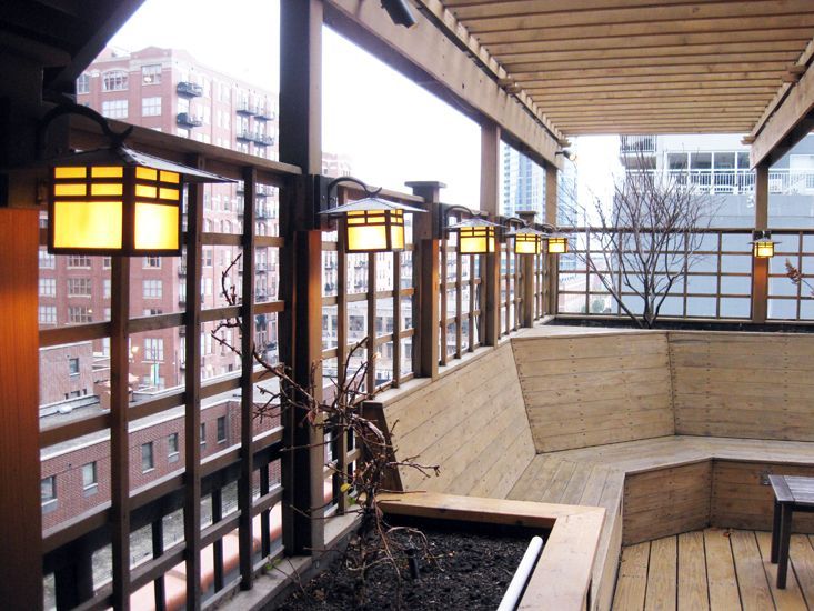 A rooftop deck featuring wooden benches, a trellis with glowing square lanterns, and a small potted tree.