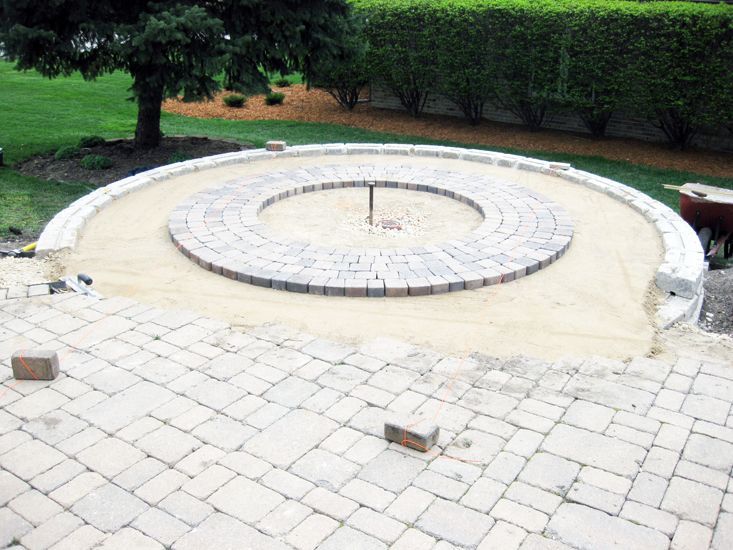 A circular brick patio under construction in a grassy yard, featuring a centered fire pit base and surrounding pavers.