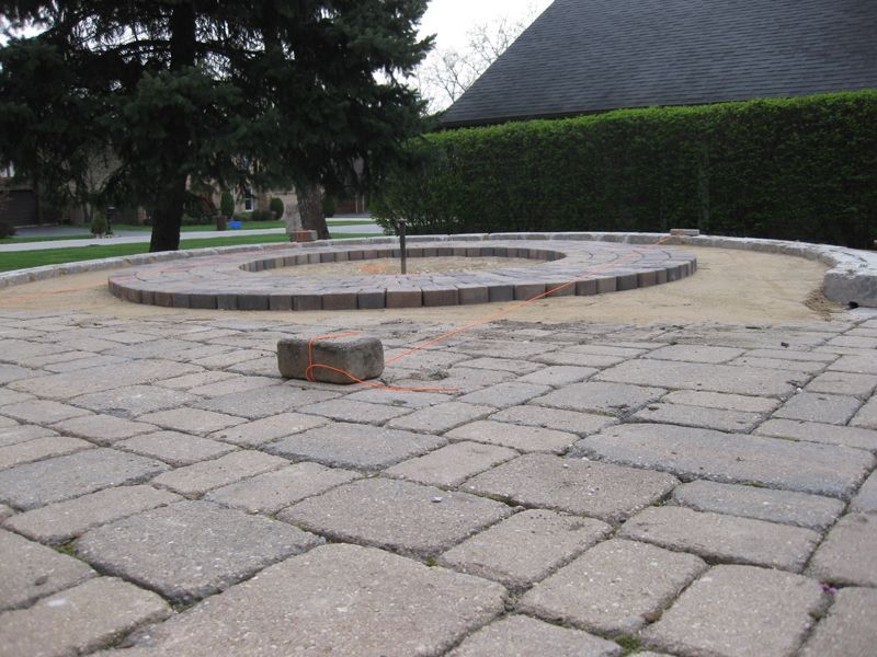 A backyard patio under construction, showing a circular stone feature set in sand on a paved ground surface.