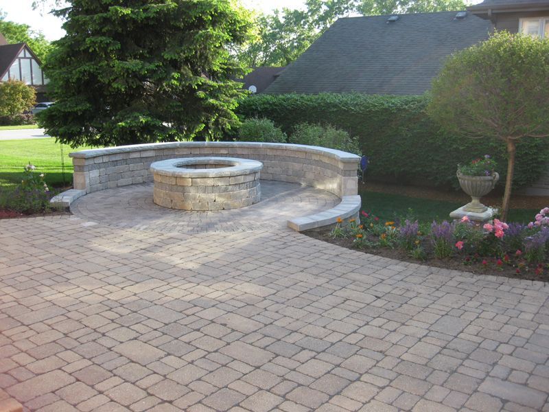 A paved outdoor patio with a circular stone fire pit and a curved stone seating wall, surrounded by trees and flowers.