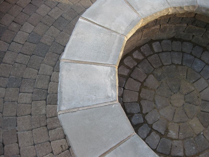 A close-up of a circular stone fire pit border made of light gray concrete blocks, surrounded by charcoal cobblestones.
